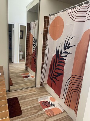 Un baño con cortina de ducha estampada con hojas tropicales, piso de madera y un espejo que refleja la habitación.