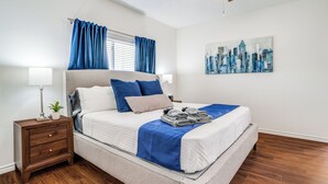 A neatly made bed with a blue and white bedspread, a wooden nightstand with drawers, a small potted plant, and a cityscape painting on the wall.