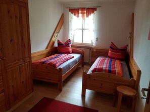 Une chambre avec deux lits en bois, une fenêtre avec des rideaux, une armoire en bois et un tapis rouge.