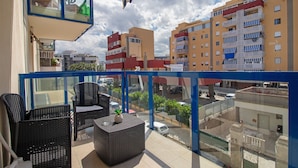 Un balcón con mobiliario de mimbre, baranda de vidrio y vista de edificios circundantes.