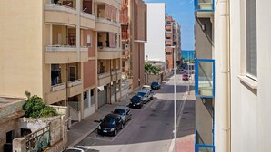 Un edificio residencial de varios pisos con balcones, una calle con coches estacionados y un cielo despejado.