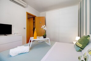 Une chambre d’hôtel avec un lit, une télévision, une commode blanche, une petite table avec une plante et une porte donnant sur une autre pièce.