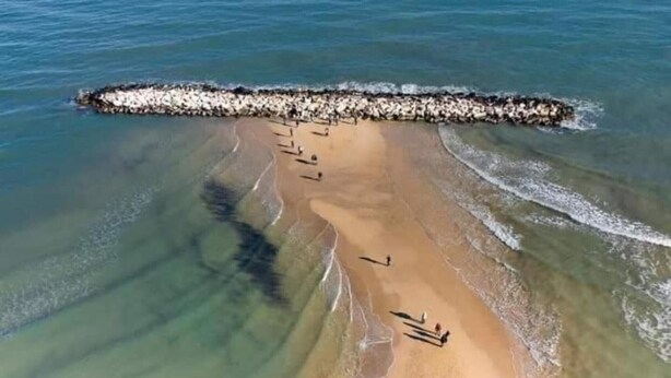 Vista aérea de una playa con un rompeolas rocoso, agua cristalina y algunas personas.