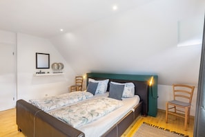 Un dormitorio con cama, dos sillas, una mesita de noche, un estante de pared con adornos y una lámpara en el techo.