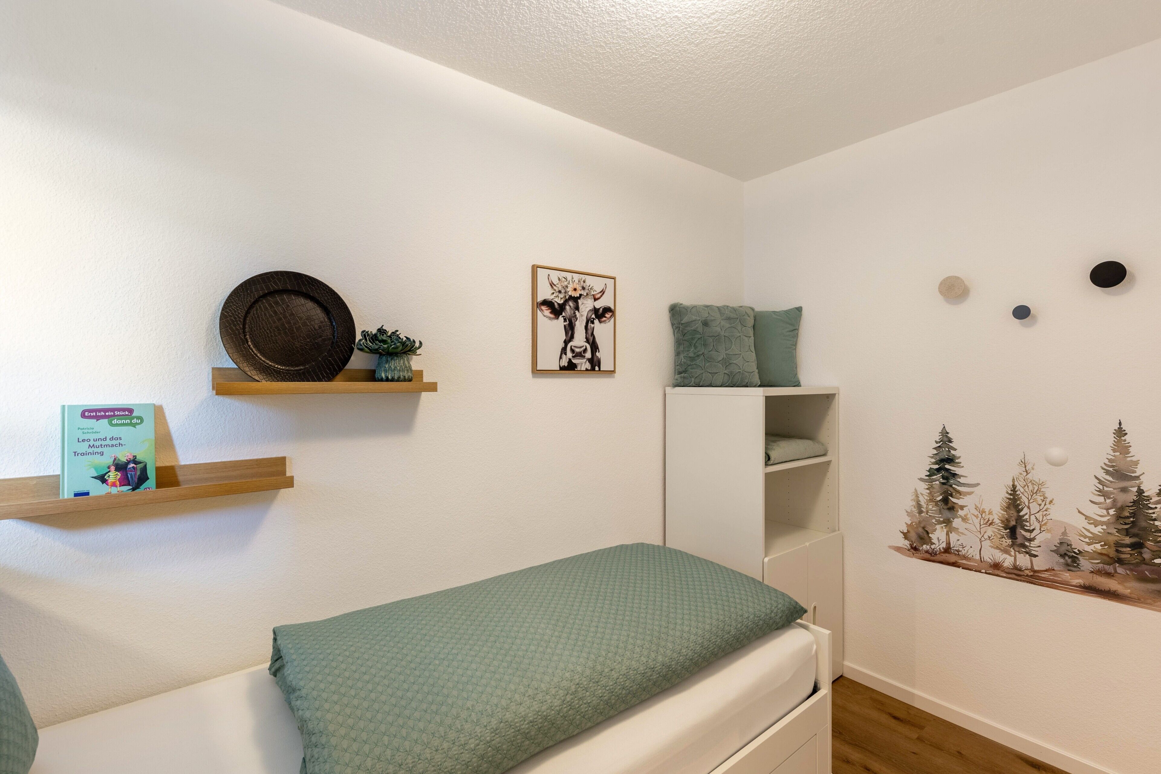 Un dormitorio con cama, repisa con un libro, decoración en la pared y un mueble pequeño.