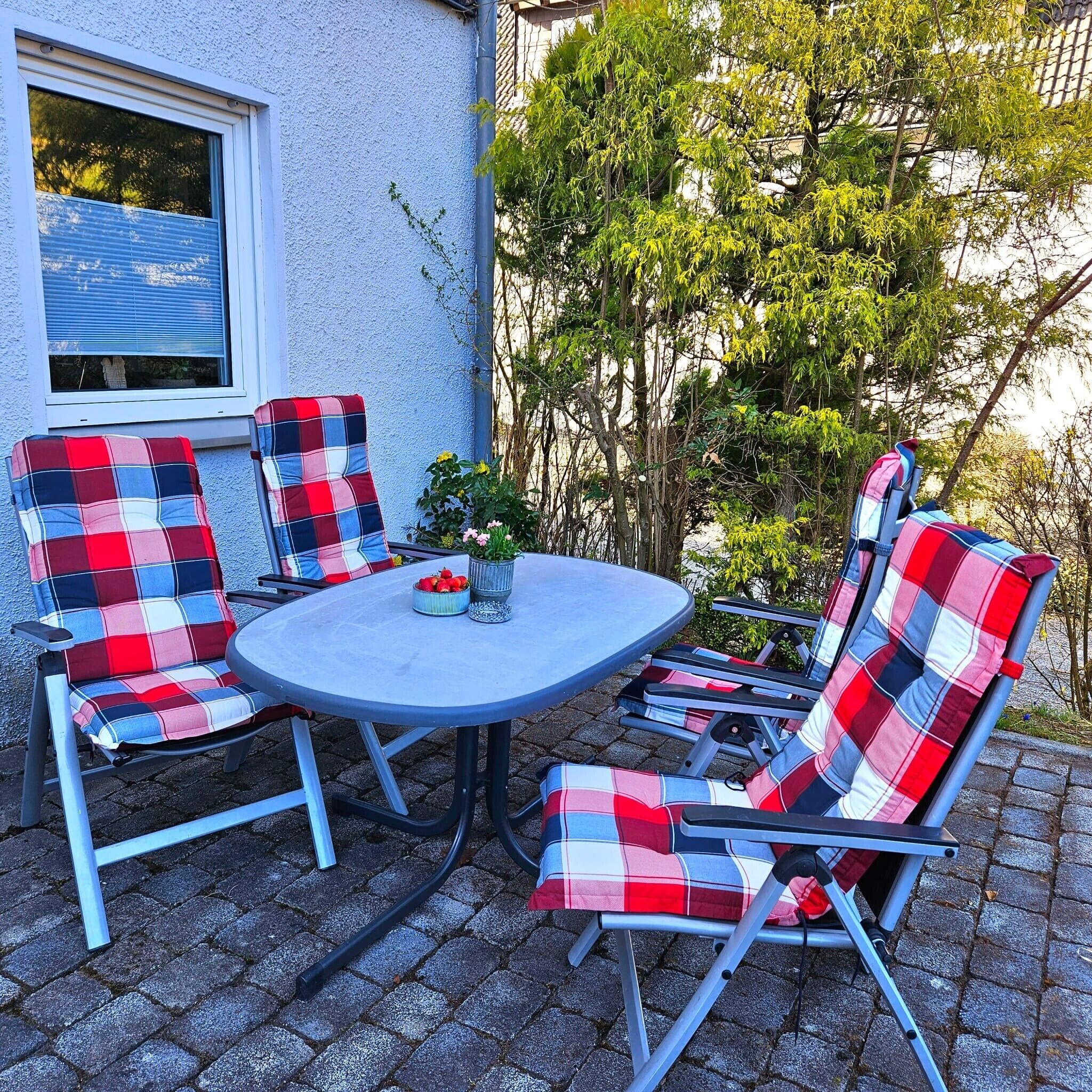 Une terrasse extérieure avec une table ronde et quatre chaises, chacune agrémentée d’un coussin à carreaux.
