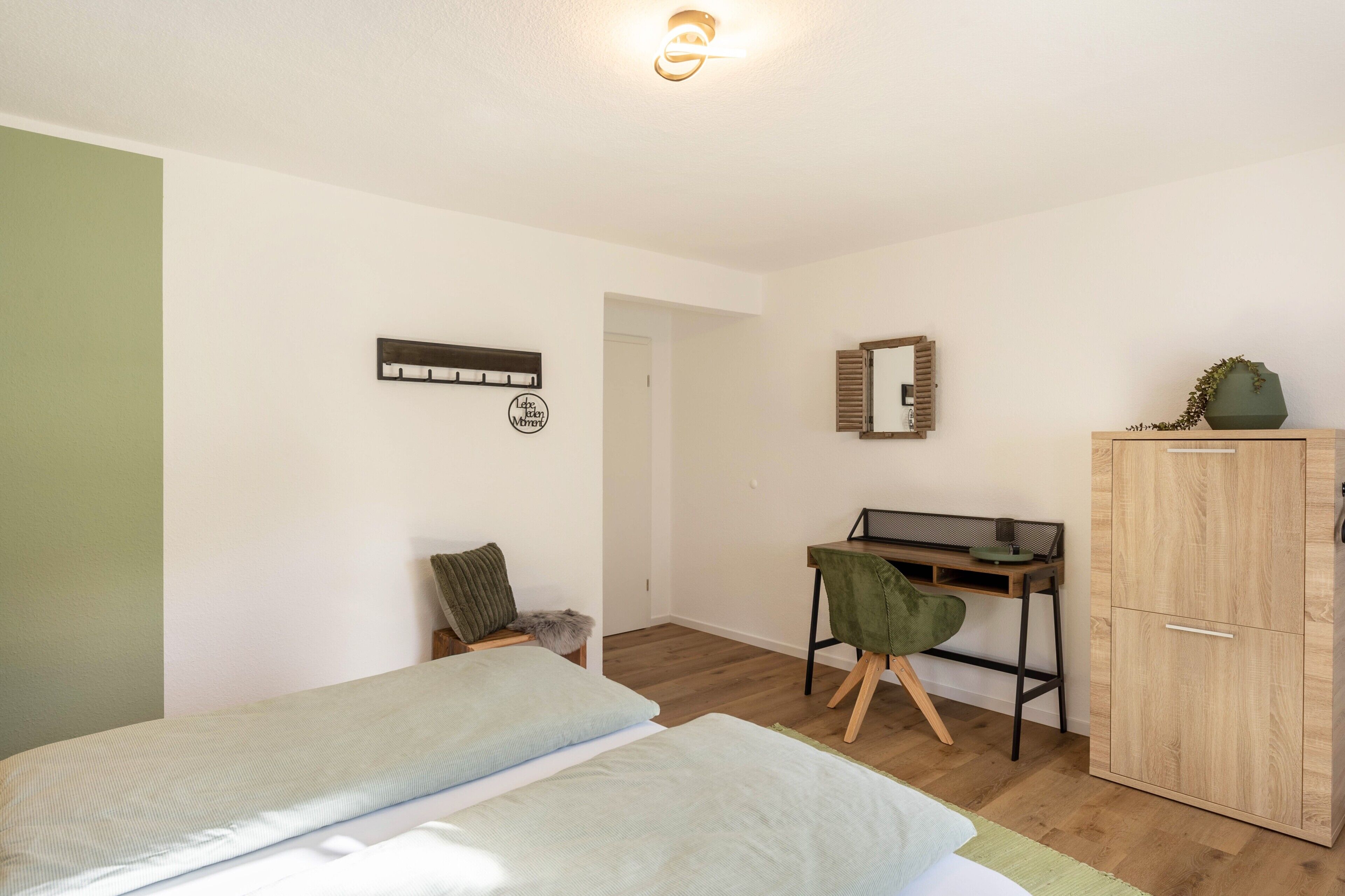 Un dormitorio con cama, escritorio, silla, armario y espejo adosado a la pared.