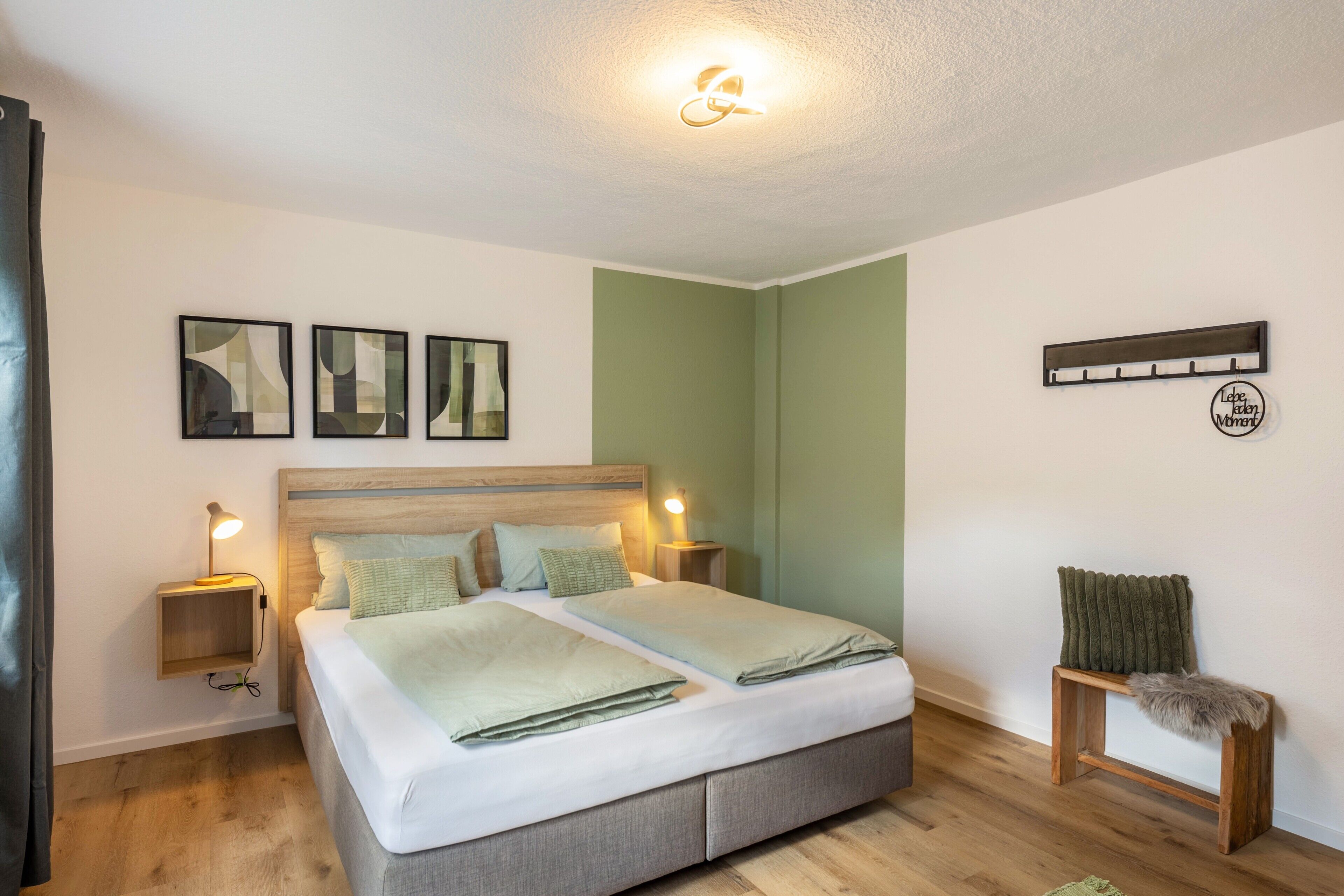 A bedroom with a double bed, bedside lamps, a small wooden bench, and abstract wall art.