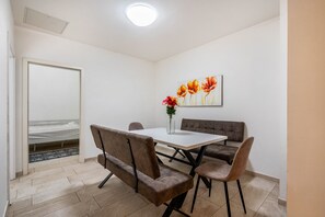 A room with a white table, four chairs, a sofa, a painting of flowers, and a bed in the background.