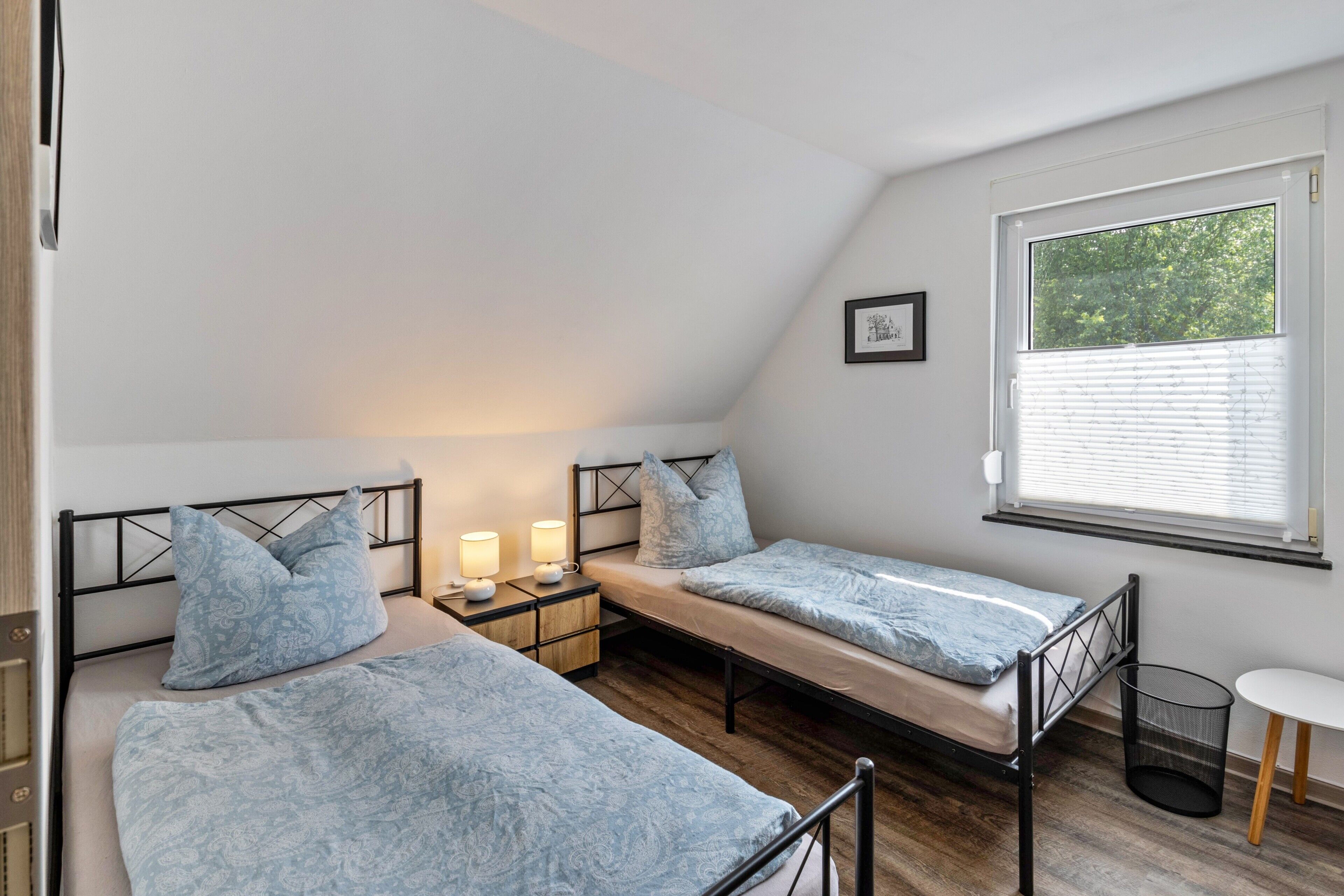A bedroom with two beds, a window with blinds, a bedside table with a lamp, and a small round table.