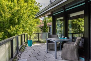 En balkon med et bord og stole, en potteplante samt udsigt til træer og et hus.