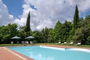 A swimming pool with lounge chairs and umbrellas, surrounded by trees and a clear sky.