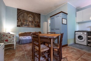 A room with a dining table, chairs, a sofa, a washing machine, and a large painting on the wall. - Apartment 'Rosaspina - 7' with Shared Pool, Shared Terrace and Air Conditioning (Capalbio)