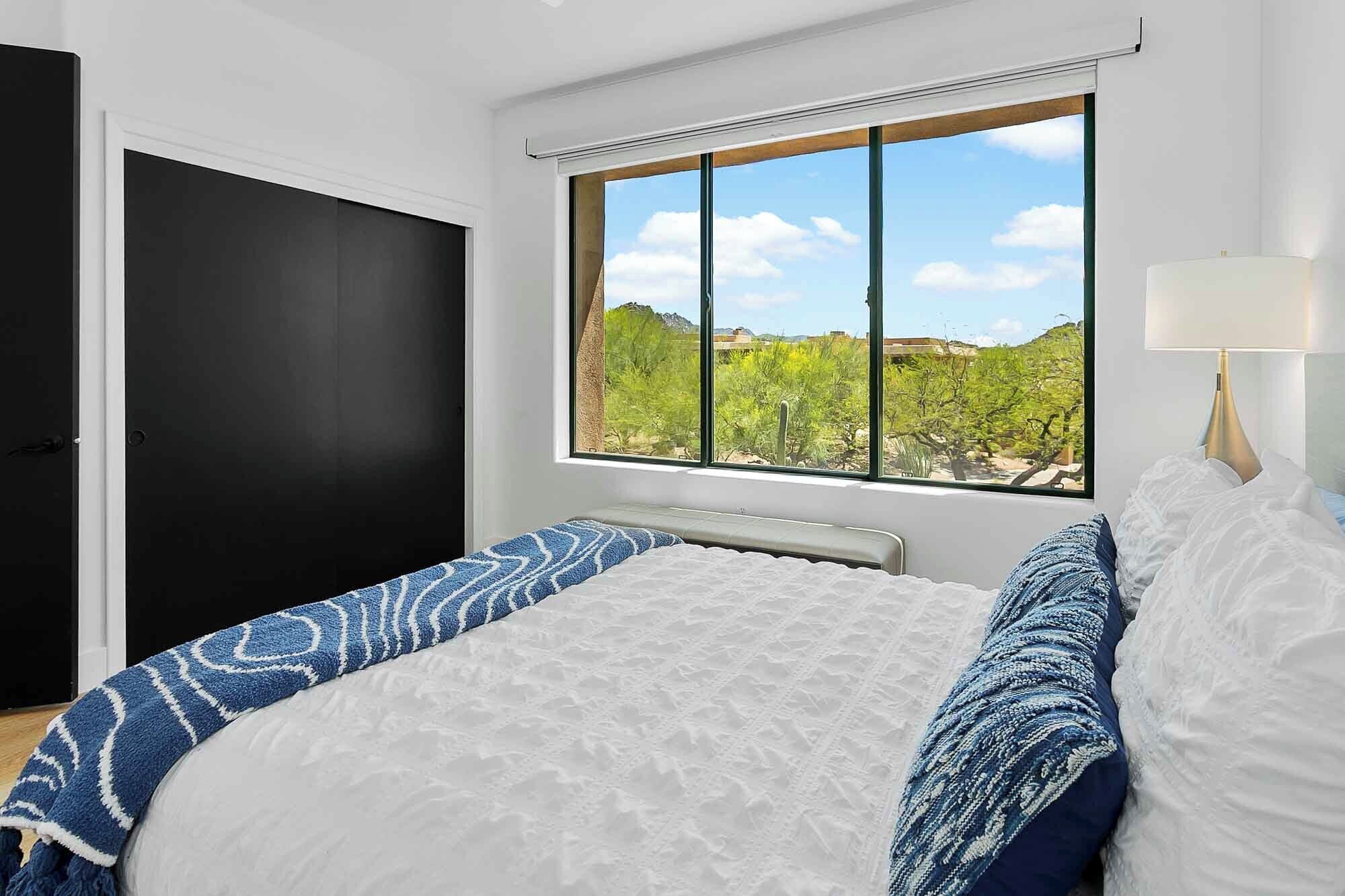 Une chambre à coucher avec un lit, une fenêtre donnant sur un paysage verdoyant, une porte noire et une lampe de chevet.