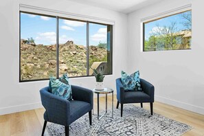 A room with two armchairs, a small table, and a patterned rug. Large windows offer views of rocky terrain and sparse vegetation.