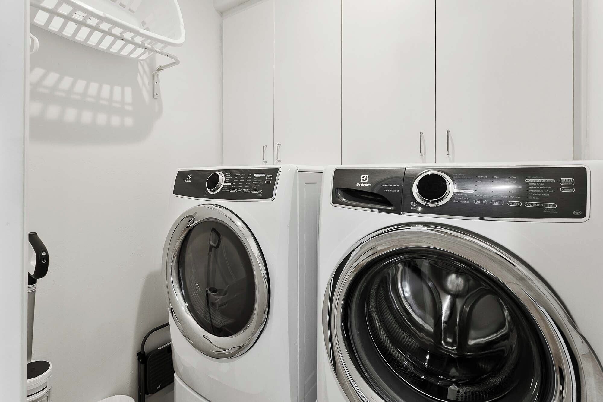 Two front-loading washing machines in a laundry room with a white wall and a metal drying rack above.