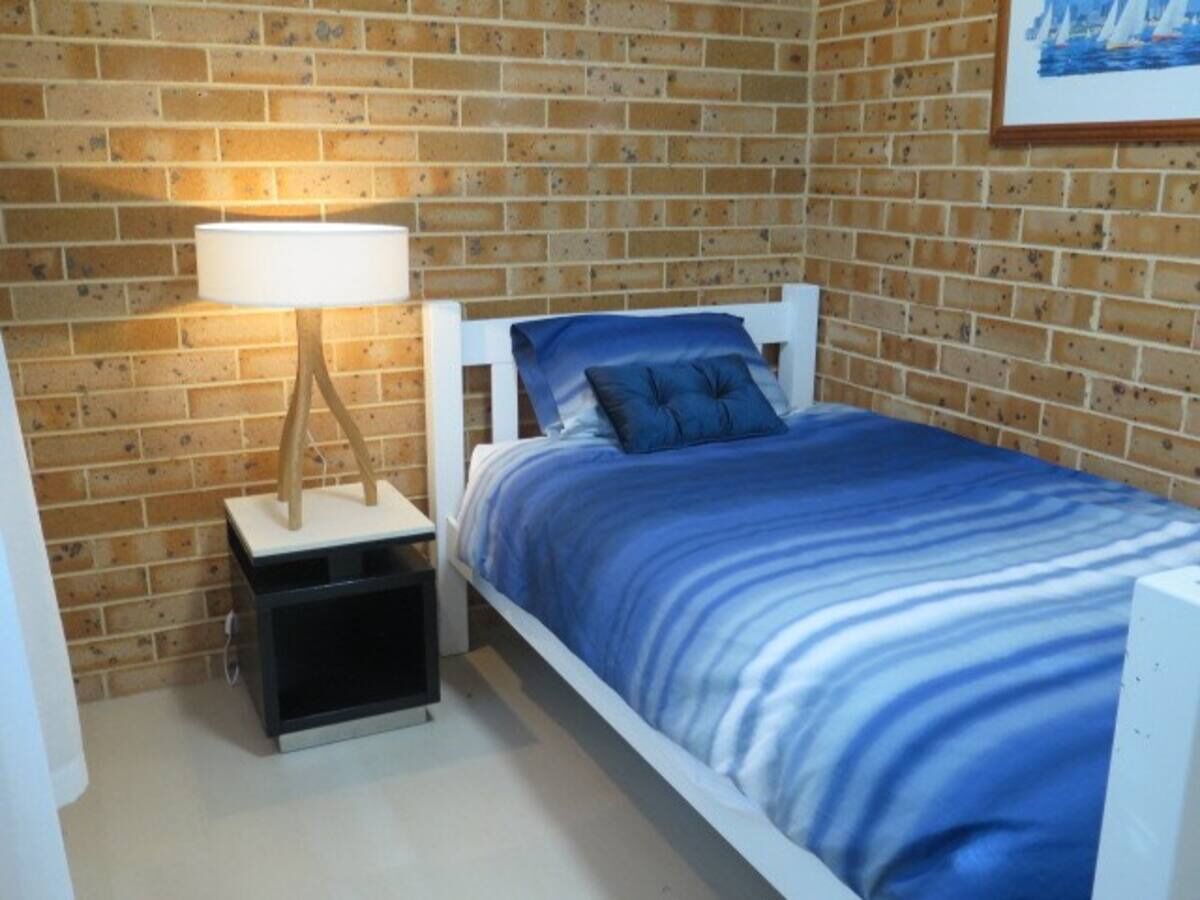 A single bed with a white frame and a blue-striped bedspread, a bedside table with a lamp, and a brick wall background.