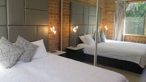 A hotel room with two beds, a brick wall, a large mirror, and a window with curtains. - Spacious 3 bedroom beach apartment. (Mona Vale)