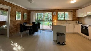 A modern kitchen with white cabinets, a built-in oven, and a dining area with a table and chairs. - Spacious 3 bedroom beach apartment. (Mona Vale)