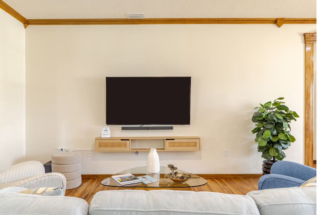 A living room with a flat-screen TV mounted on the wall, a glass coffee table, a potted plant, and a sofa.
