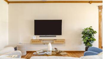 A living room with a flat-screen TV mounted on the wall, a glass coffee table, a potted plant, and a sofa.