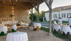 Un área de comedor al aire libre con mesas dispuestas bajo una pérgola de madera, rodeada de muros de piedra y una zona ajardinada.