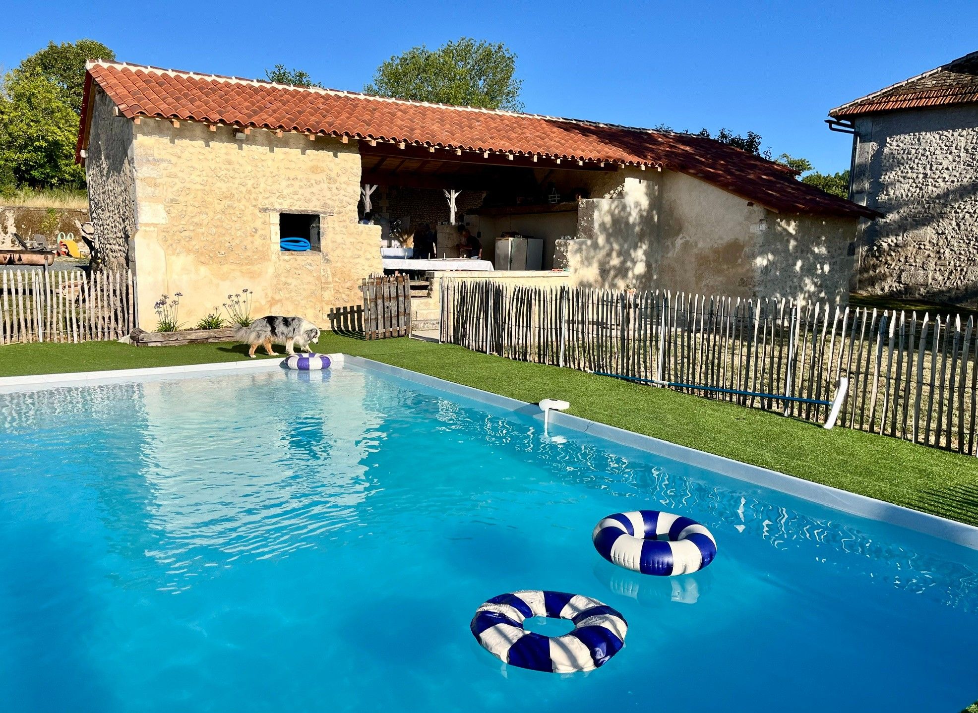 A swimming pool with two inflatable rings, a stone building with a red-tiled roof, and a wooden fence.