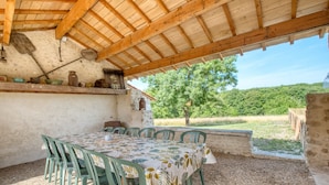 Een terras buiten met een tafel en stoelen, een houten plafond en uitzicht op een groen landschap.