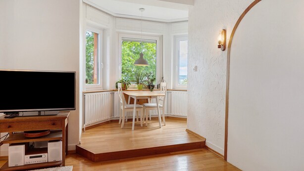 A room with a television on a wooden stand, a dining table with chairs, a large window with a view of greenery, and a white door. - Apartment 'Rotes Haus' with Mountain View, Private Terrace and Wi-Fi (Konstanz)