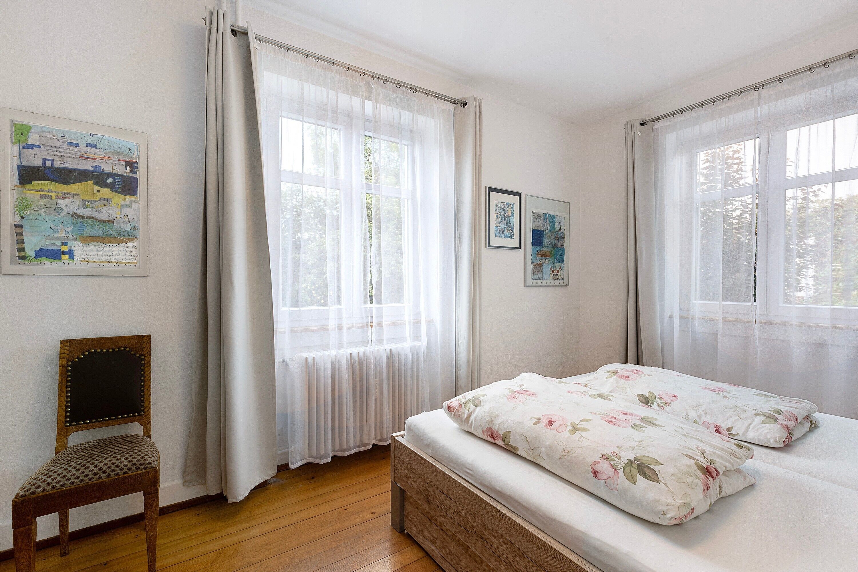 A bedroom with a wooden bed, a chair, and two windows with sheer curtains.