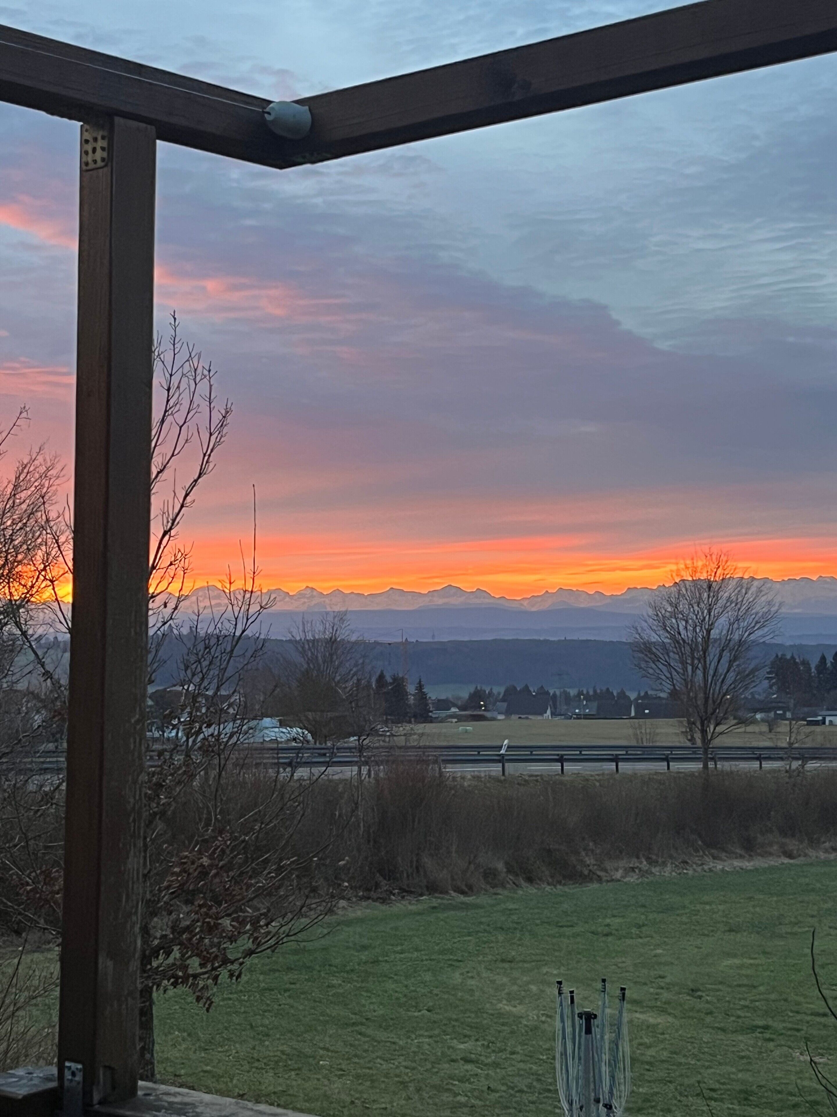 Sonnenuntergangblick von einer überdachten Außenbereitschaft mit Holzkonstruktion, einem Grasfeld und fernen Bergen.