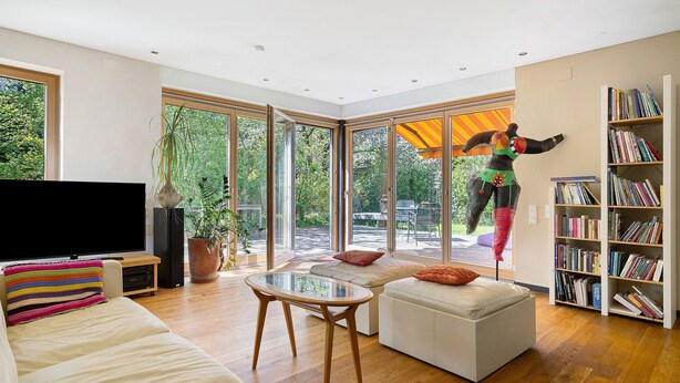 A modern living room with a large window, a flat-screen TV, a bookshelf, and a sofa. - Loft with Fireplace, Whirlpool, Private Terrace, Garden, and Wi-Fi (Lustenau)