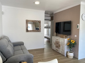 A living room with a grey sofa, a mounted TV, a wooden-framed mirror, and a potted plant with yellow flowers.