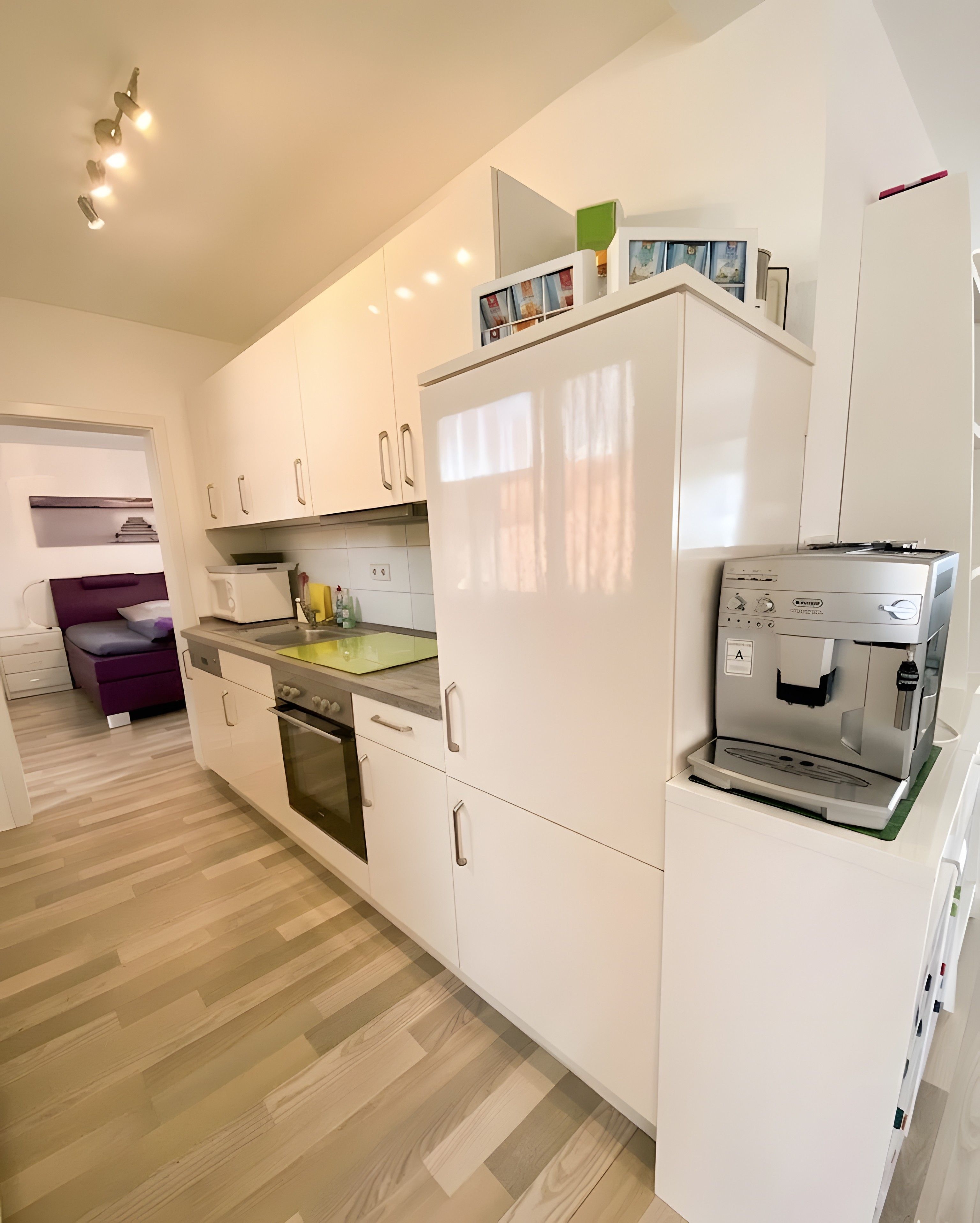 A modern kitchen with white cabinets, a built-in oven, a coffee machine, and a dining area with a purple chair.