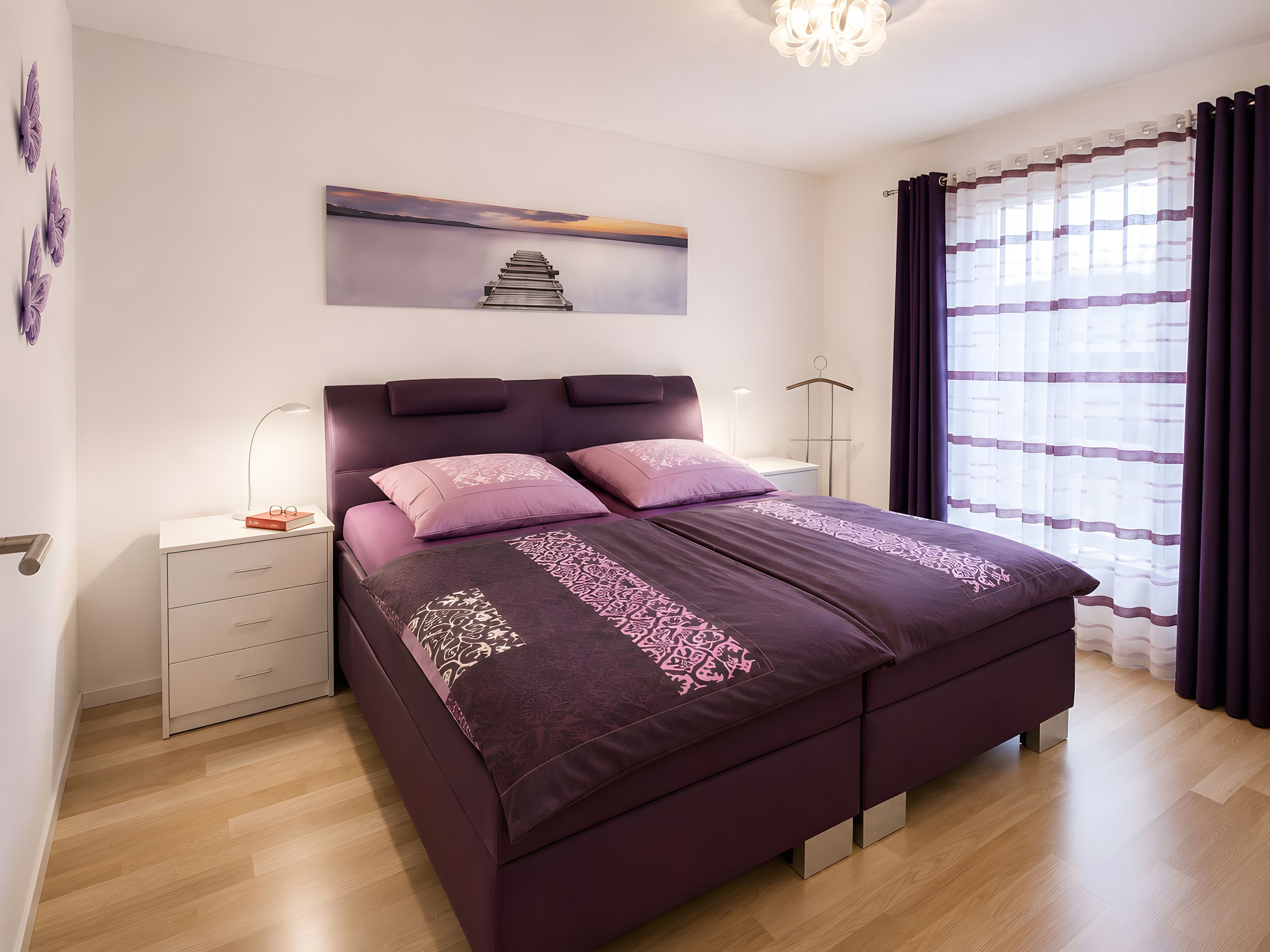 A modern bedroom with a large bed, bedside tables, a nightstand, a hanging light, and a view of the outdoors through a window with curtains.