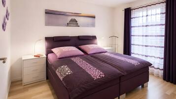 A modern bedroom with a large bed, bedside tables, a nightstand, a hanging light, and a view of the outdoors through a window with curtains.