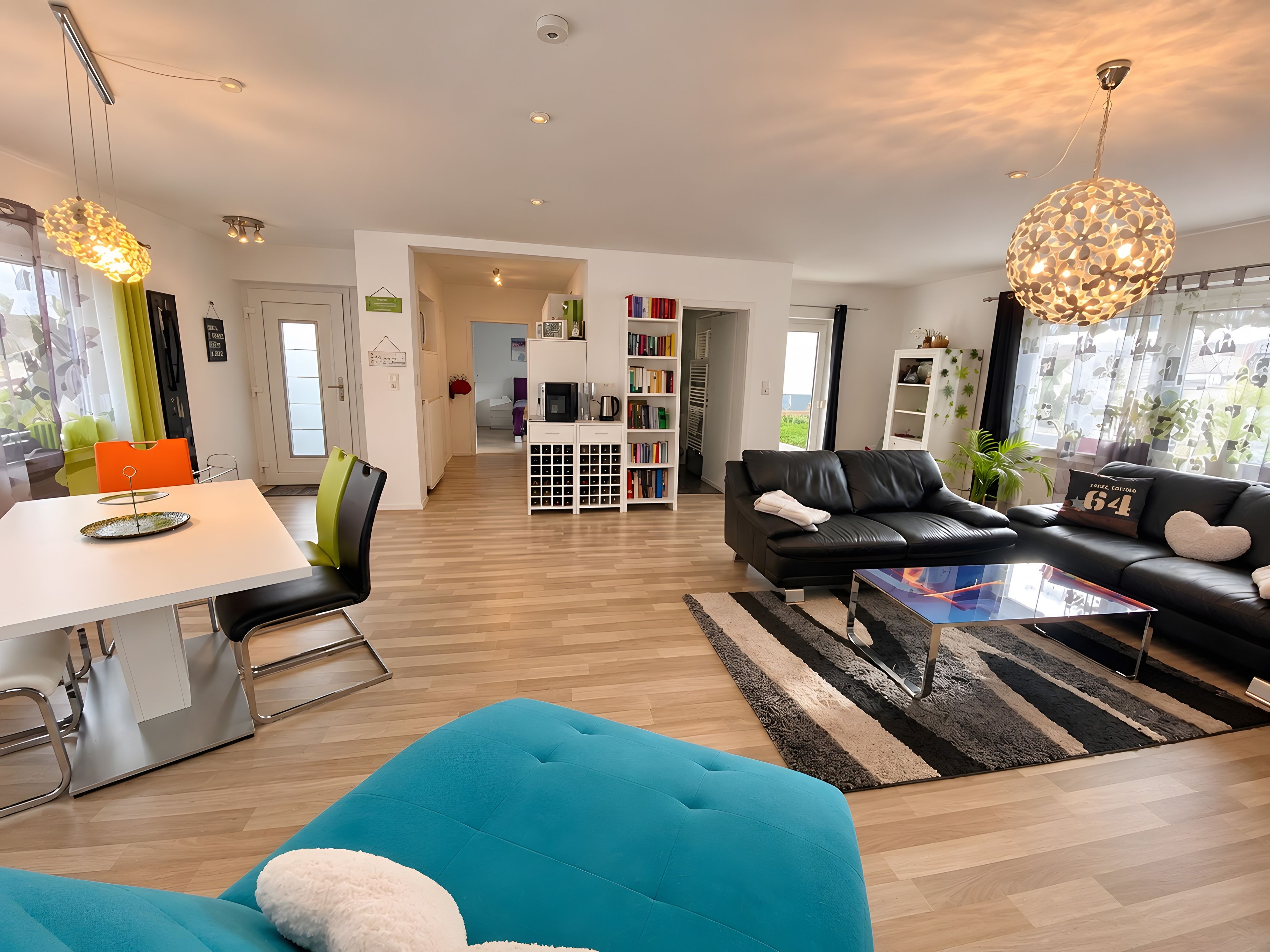 A modern living room with a black leather sofa, a teal ottoman, and a glass coffee table. There is a white bookshelf filled with books and a white cabinet with a microwave.