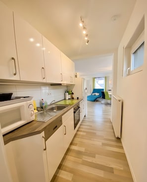 A modern kitchen with white cabinets, a microwave, and a sink. There is a dining area with a blue chair and a window.