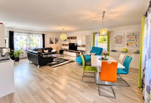 A modern living room with a sofa, a dining table with chairs, a kitchen area, and a large window with curtains.