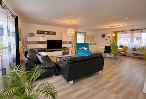 A modern living room with a black sofa, a flat-screen TV, and a dining area with colorful chairs.