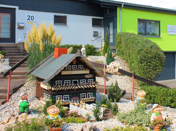 A model house with a sloped roof, surrounded by rocks and plants, with a small statue of a person in front.
