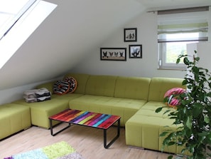 A green sofa, a colorful coffee table, and a plant in a room with a sloped ceiling and a window.