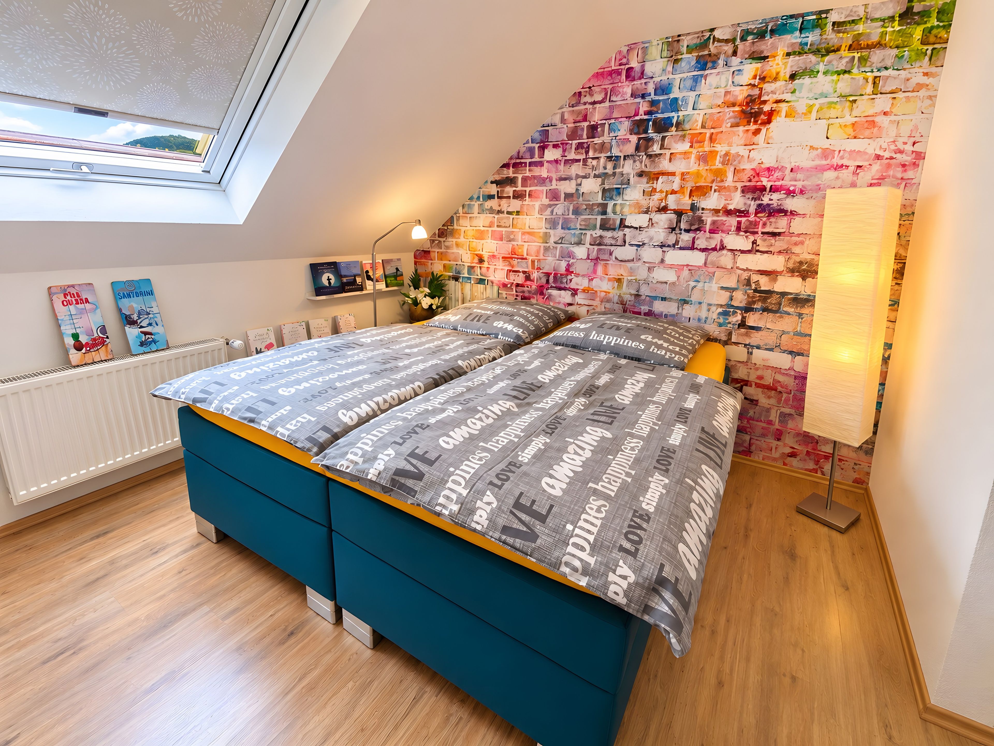 Ein Schlafzimmer mit schräg stehender Decke, einer farbenfrohen Ziegelwand, einem Bett mit gemusterten Betttextilien, einem Heizkörper und einem Fenster mit Aussicht.