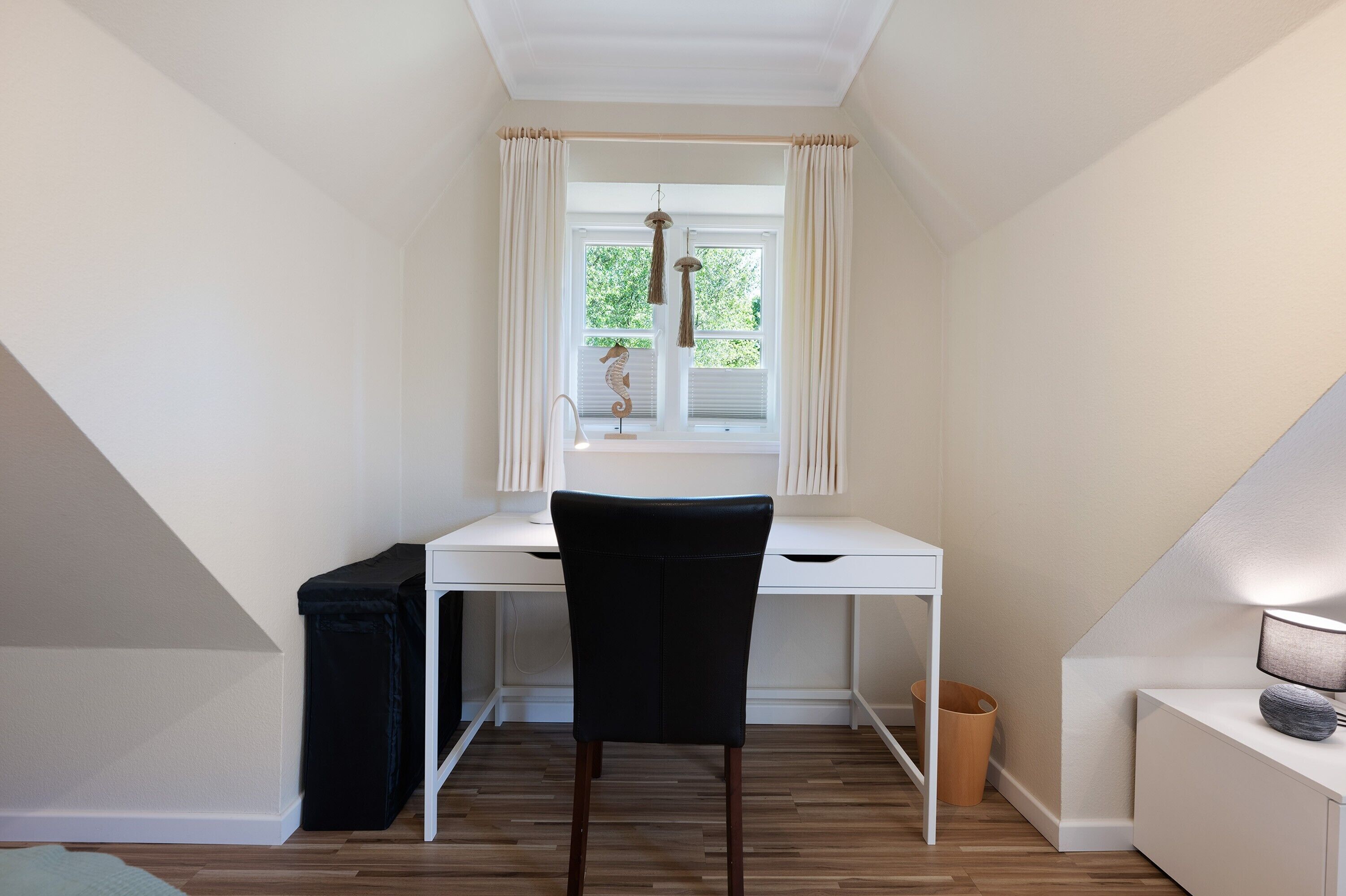A small, white desk with a black chair, a window with curtains, a hanging ornament, and a lamp on a side table.