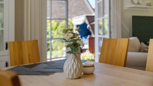 A dining table with a white vase and a small bowl, wooden chairs, and a view of a house through large windows. - Holiday Home 'Huus Alex' with Private Terrace, Private Garden and Wi-Fi (St. Peter-Ording Dorf)