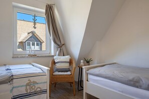 A bedroom with two beds, a wicker chair, a small table with a plant, and a window with a view of a house.
