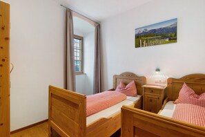 Une chambre à coucher avec deux lits en bois, une fenêtre avec des rideaux, une table de chevet avec une lampe et un tableau représentant un paysage de montagne accroché au mur.