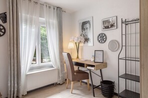 Een kamer met een bureau, stoel, radiator, raam met gordijnen en wanddecoraties, waaronder ingelijste foto's en een metalen plank.