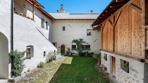 A courtyard with a stone pathway, a grassy area, and a building with wooden beams and white walls. - Apartment 'Gravetsch' with Mountain View, Shared Garden and Wi-Fi (Villandro)
