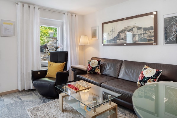 A living room with a glass coffee table, a brown leather sofa, a black armchair, and a chessboard.